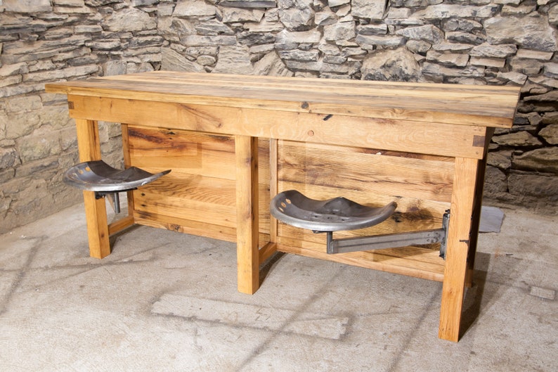 May include: A rustic wooden bar table with two metal tractor seat stools attached to the underside. The table has a natural wood finish and is made from reclaimed wood. The stools are black metal and have a simple design.
