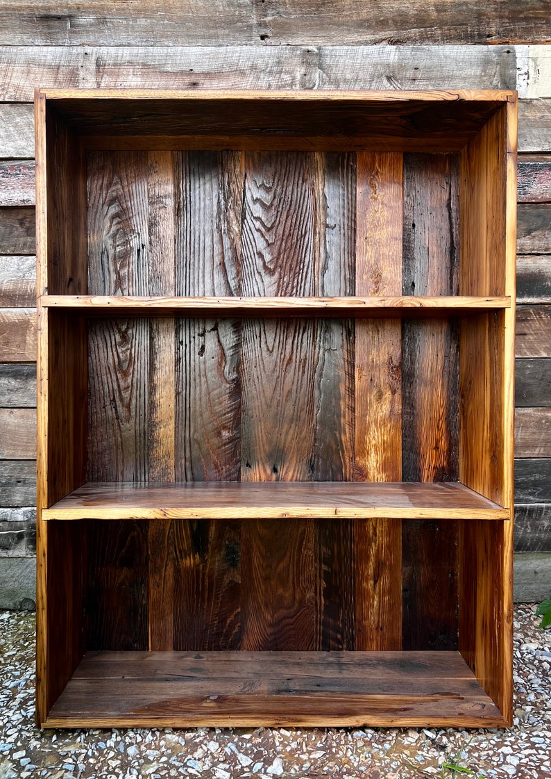 May include: A rustic wooden bookshelf with three shelves. The wood has a natural, varied grain and color, ranging from light brown to dark brown. The bookshelf is rectangular and appears to be handmade.