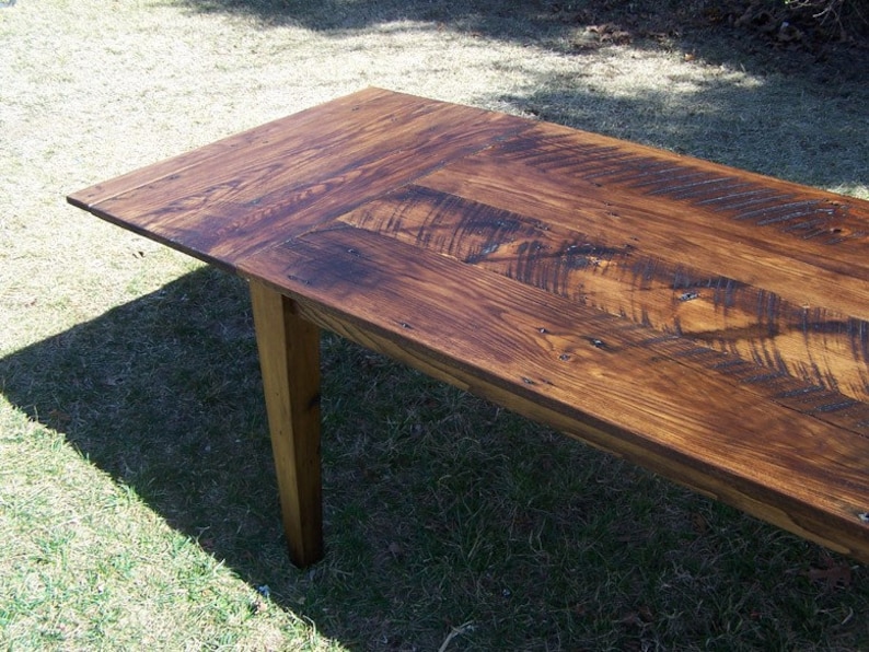 May include: A rustic wooden table with a dark brown stain. The table top is made of reclaimed wood planks with a knotty grain pattern. The table has a single visible leg made of solid wood.