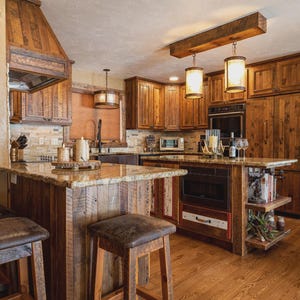May include: A rustic kitchen with wooden cabinets, a granite countertop, and a central island. Two pendant lights hang above the island. The kitchen features a wooden range hood, a refrigerator, and bar stools. The flooring is hardwood.