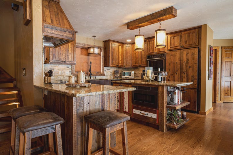 May include: A rustic kitchen with wooden cabinets, a granite countertop, and a central island. Two pendant lights hang above the island. The kitchen features a wooden range hood, a refrigerator, and bar stools. The flooring is hardwood.