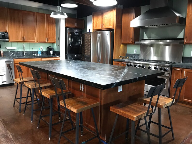 May include: A kitchen with a large island featuring a dark countertop and wooden cabinets. Several metal and wood bar stools are positioned around the island. Stainless steel appliances and pendant lights are also visible.