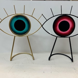 Puede incluir: Dos esculturas decorativas de ojos de metal con acabado dorado y negro. Los ojos son redondos y tienen un esquema de color turquesa y rosa.