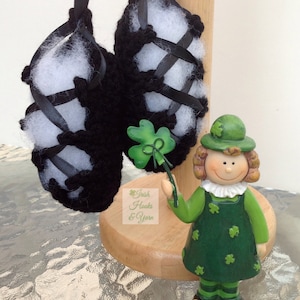 May include: Black crocheted Irish dancing shoes with white felt lining and black ribbon laces. The shoes are displayed on a wooden stand with a small green shamrock figurine holding a shamrock stem.