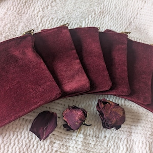 May include: A collection of six burgundy velvet pouches with gold zippers, arranged on a white, textured surface. Dried rose petals are scattered near the pouches, adding a touch of elegance to the presentation.