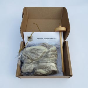May include: Open cardboard box containing a drop spindle, a package of wool roving, and a printed instruction sheet. The drop spindle is made of wood. The wool roving is a mix of white and brown fibers. The instruction sheet reads "Spinning on a Drop Spindle."