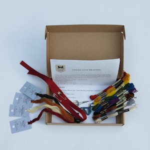 May include: A brown cardboard box open to reveal a finger loop braiding kit. The kit includes embroidery floss in various colors, a safety pin, a red ribbon, and instructions. The instructions include the text "Finger Loop Braiding".