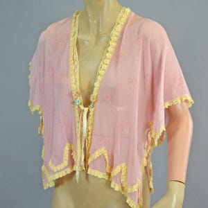 Puede incluir: Capa rosa transparente con ribete de encaje amarillo y cierre de lazo. La capa presenta un estampado floral y se exhibe en un maniquí. Los bordes están acabados con un ribete amarillo festoneado.