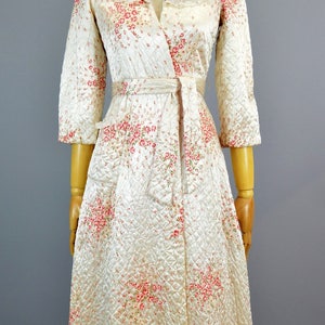 May include: A quilted robe in a cream color featuring a delicate pink and red floral pattern. This wrap-style garment includes three-quarter length sleeves, a wide collar with lace trim, and a matching fabric tie belt at the waist.