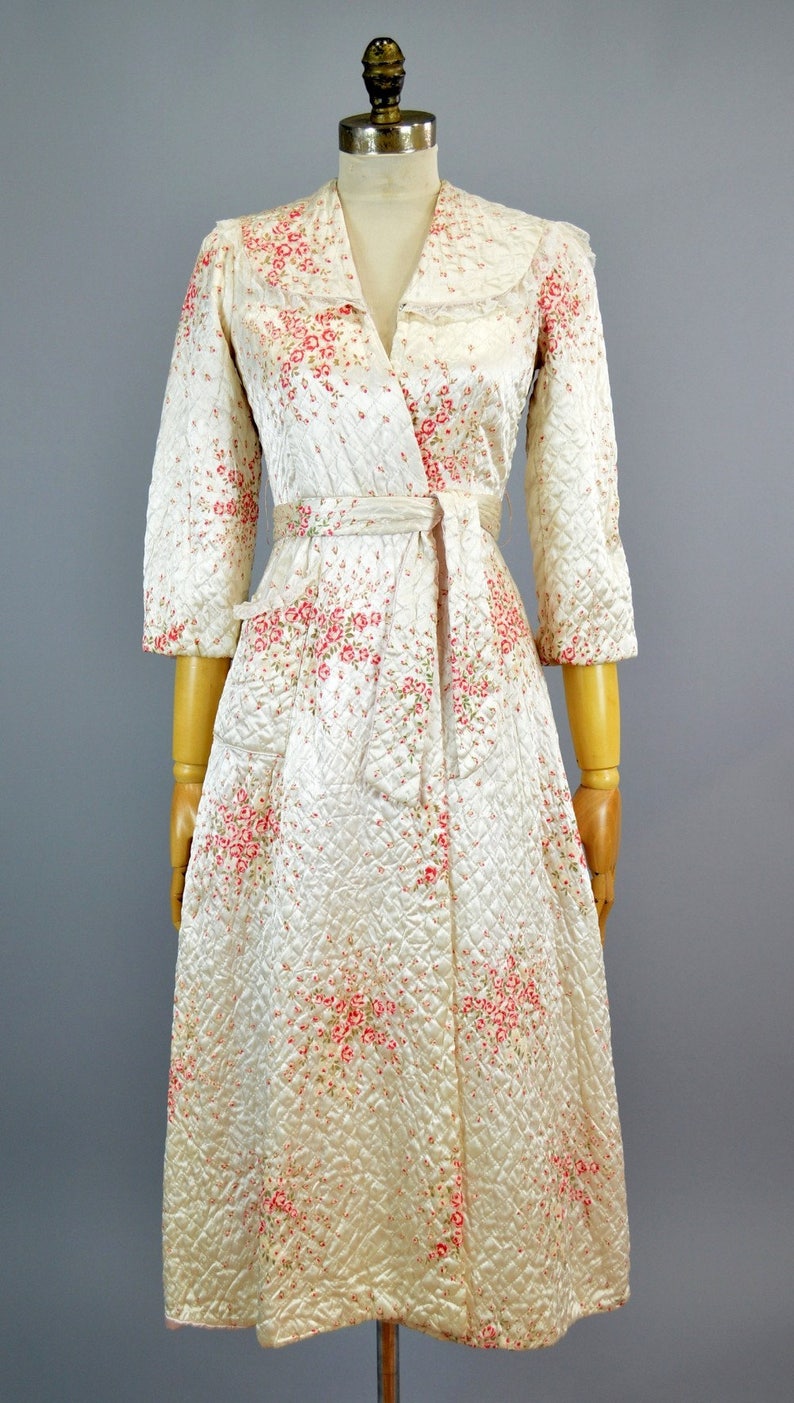 May include: A quilted robe in a cream color featuring a delicate pink and red floral pattern. This wrap-style garment includes three-quarter length sleeves, a wide collar with lace trim, and a matching fabric tie belt at the waist.