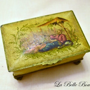 Può includere: Una scatola di legno vintage con una scena dipinta di una persona seduta sotto un tetto di paglia. La scatola ha un aspetto usurato e antico con una finitura ingiallita e un effetto di vernice screpolata.