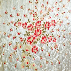 May include: A cream-colored quilted fabric featuring a diamond-stitched pattern and a central floral print. The design displays clusters of red and pink roses with green leaves, interspersed with smaller rosebuds across the entire surface.