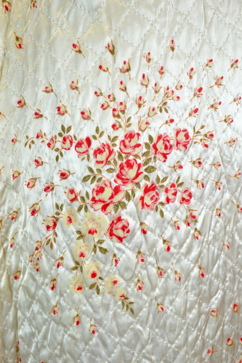 May include: A cream-colored quilted fabric featuring a diamond-stitched pattern and a central floral print. The design displays clusters of red and pink roses with green leaves, interspersed with smaller rosebuds across the entire surface.