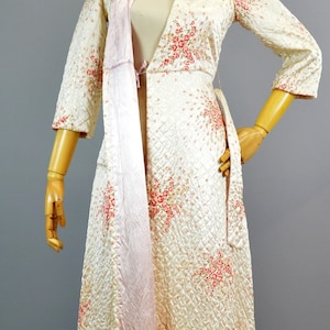 May include: A long, quilted robe in a cream-colored fabric featuring a delicate pink and red floral pattern. The garment has three-quarter length sleeves, a wide collar, and a smooth, light pink satin lining visible along the front opening.