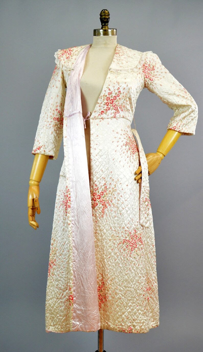 May include: A long, quilted robe in a cream-colored fabric featuring a delicate pink and red floral pattern. The garment has three-quarter length sleeves, a wide collar, and a smooth, light pink satin lining visible along the front opening.