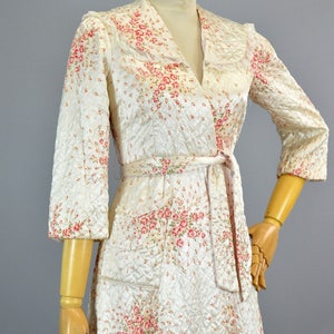 May include: A quilted robe featuring a white base with a pink and green floral pattern, a shawl collar, three-quarter length sleeves, and a matching tie belt at the waist. The garment has a diamond-stitched texture and two front patch pockets.