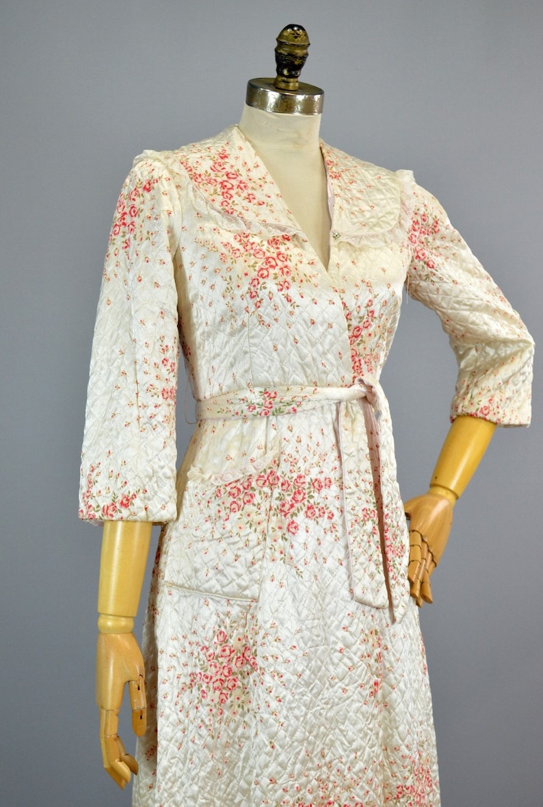 May include: A quilted robe featuring a white base with a pink and green floral pattern, a shawl collar, three-quarter length sleeves, and a matching tie belt at the waist. The garment has a diamond-stitched texture and two front patch pockets.