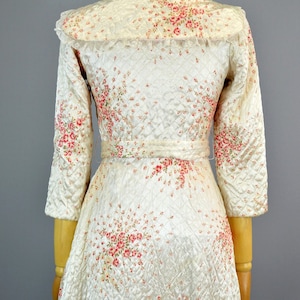 May include: A cream-colored quilted robe featuring a delicate pink and red floral print, three-quarter length sleeves, a wide collar with lace trim, and a matching fabric belt tied at the waist, displayed on a mannequin from the back view.