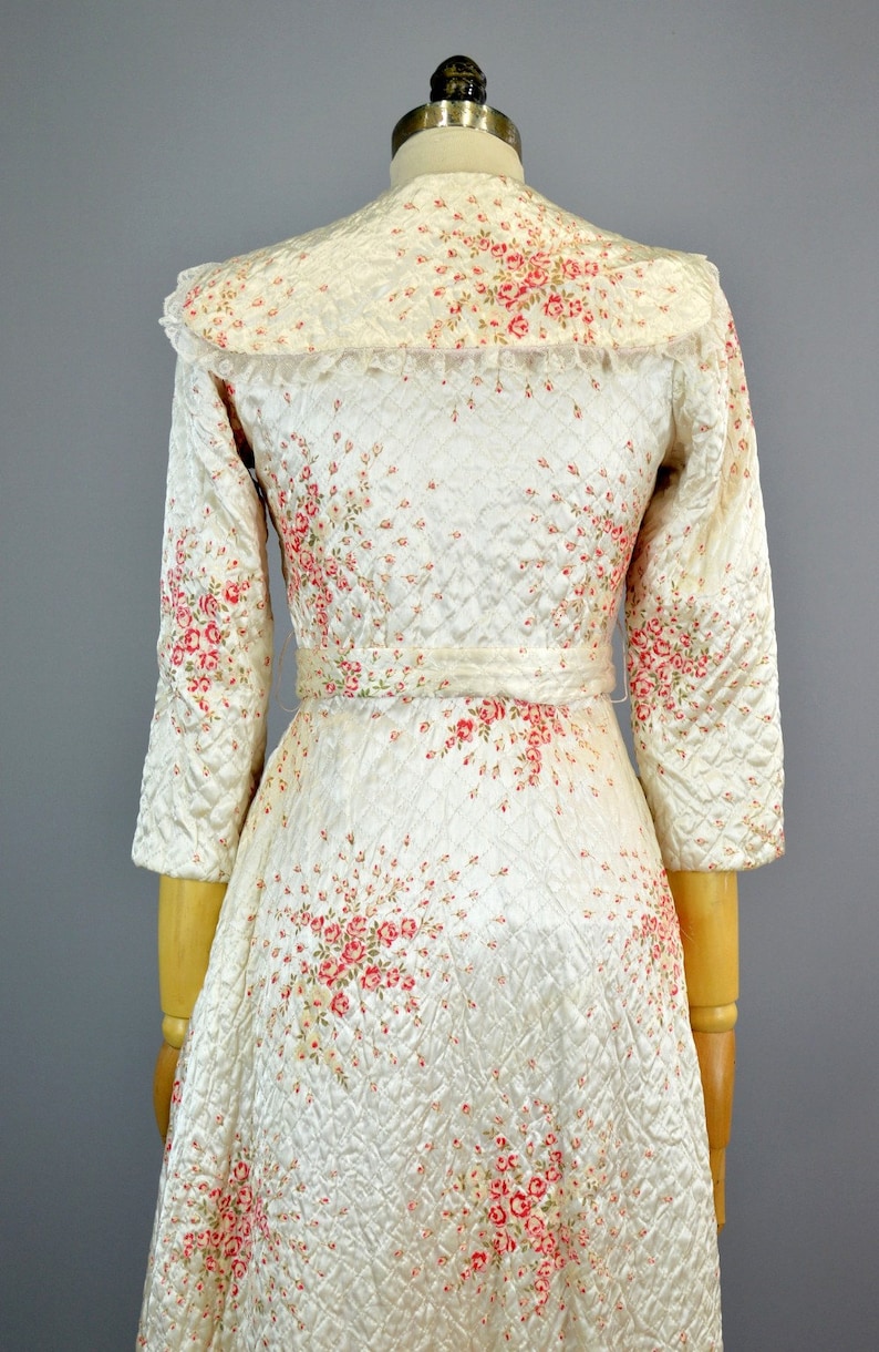 May include: A cream-colored quilted robe featuring a delicate pink and red floral print, three-quarter length sleeves, a wide collar with lace trim, and a matching fabric belt tied at the waist, displayed on a mannequin from the back view.