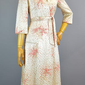 May include: A cream-colored quilted robe featuring a delicate pink and green floral pattern. This garment has three-quarter length sleeves, a shawl collar, and a matching fabric tie belt at the waist. The quilted texture covers the entire piece.