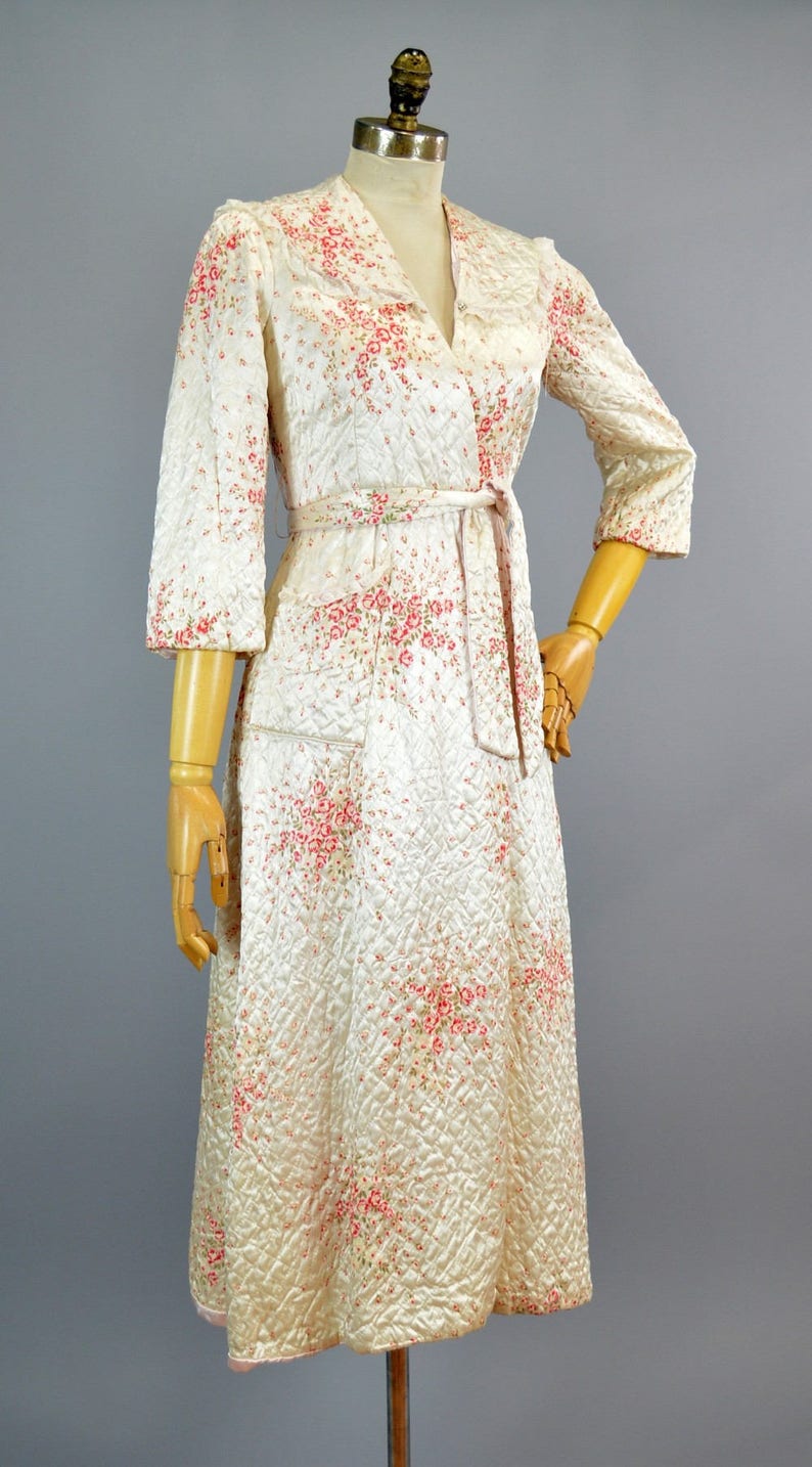 May include: A cream-colored quilted robe featuring a delicate pink and green floral pattern. This garment has three-quarter length sleeves, a shawl collar, and a matching fabric tie belt at the waist. The quilted texture covers the entire piece.