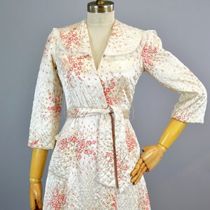 May include: A quilted robe in a cream color featuring a pink and green floral pattern. The garment has three-quarter length sleeves, a wide collar with lace trim, and a matching fabric tie belt at the waist. The fabric has a diamond-stitched texture.