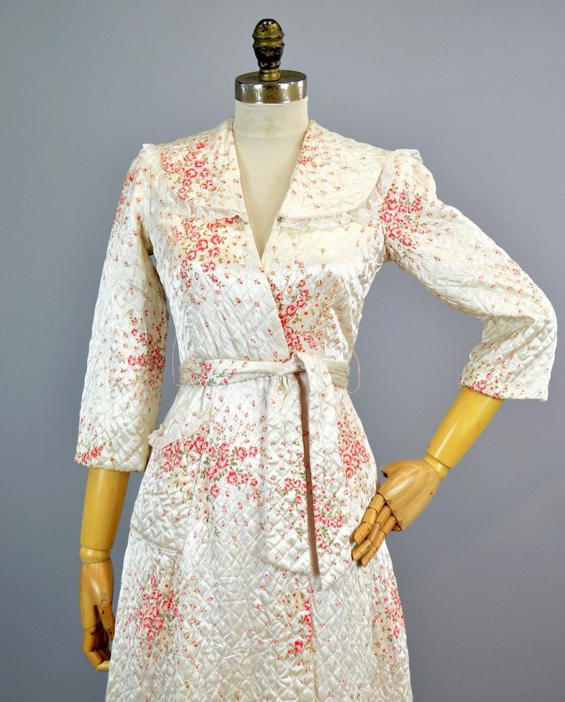 May include: A quilted robe in a cream color featuring a pink and green floral pattern. The garment has three-quarter length sleeves, a wide collar with lace trim, and a matching fabric tie belt at the waist. The fabric has a diamond-stitched texture.