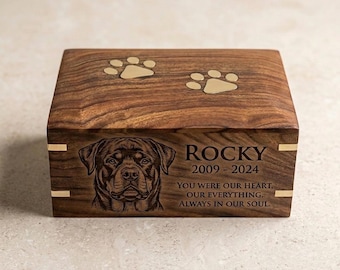 May include: A wooden pet urn with a detailed engraving of a Rottweiler's face. The top features two paw prints. The name "Rocky" and dates 2009-2024 are inscribed, along with a heartfelt message. Gold accents adorn the corners.