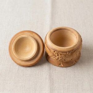 May include: A small, round wooden box with a lid, both in a light brown color. The box has carved floral designs around the sides. The lid is off to the side, revealing the interior of the box.