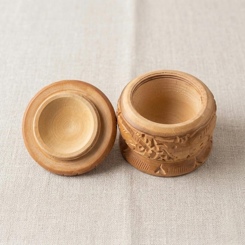 May include: A small, round wooden box with a lid, both in a light brown color. The box has carved floral designs around the sides. The lid is off to the side, revealing the interior of the box.