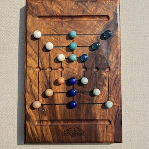 May include: A wooden board game with a dark wood grain finish. The game board features a grid of lines and holes, with various colored marbles in play. The text "WOOD & MARBLE GAMES" is printed on the board.