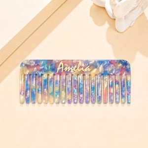 May include: A rectangular hair comb featuring a multi-colored marbled pattern with shades of blue, pink, and yellow. The name Amelia is printed in gold script lettering across the center of the comb, which has evenly spaced teeth.