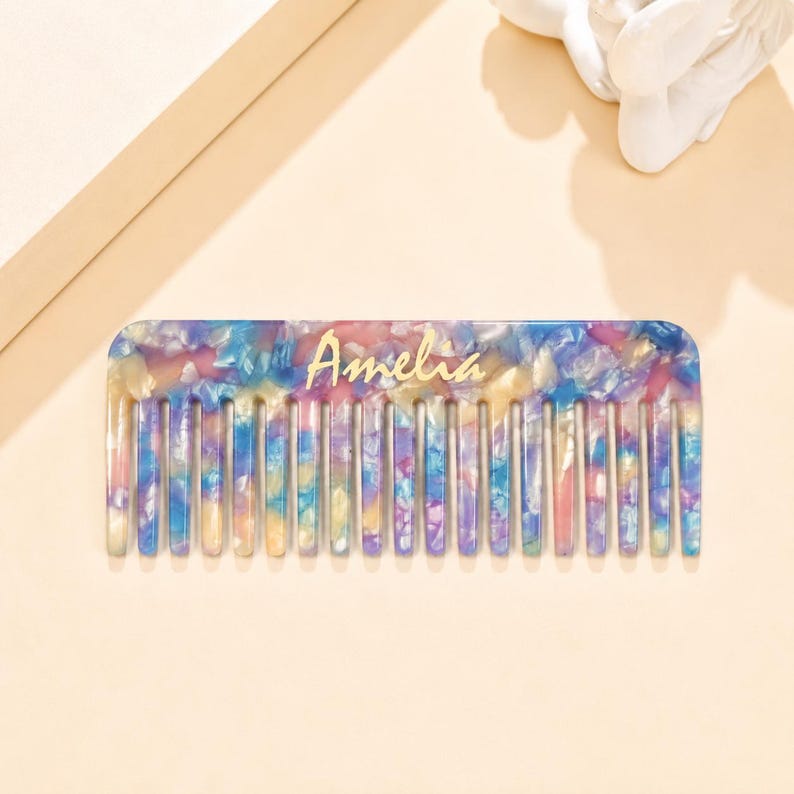 May include: A rectangular hair comb featuring a multi-colored marbled pattern with shades of blue, pink, and yellow. The name Amelia is printed in gold script lettering across the center of the comb, which has evenly spaced teeth.