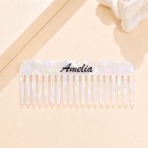 May include: A rectangular hair comb with a pearlescent white finish featuring iridescent flecks throughout the material. The name Amelia is printed in a black cursive script across the center of the comb, which has fourteen evenly spaced teeth.