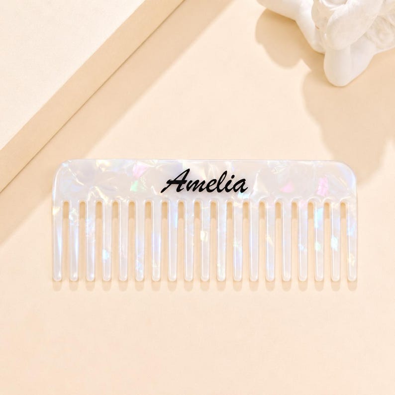 May include: A rectangular hair comb with a pearlescent white finish featuring iridescent flecks throughout the material. The name Amelia is printed in a black cursive script across the center of the comb, which has fourteen evenly spaced teeth.