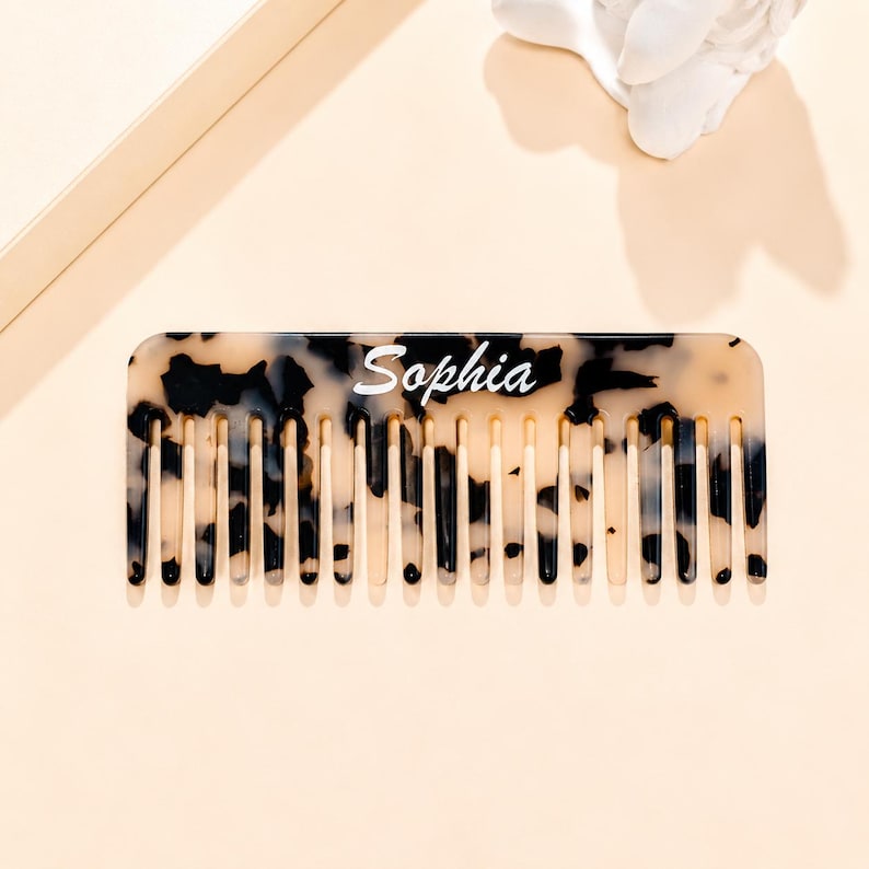 May include: A rectangular hair comb featuring a beige and black tortoiseshell pattern. The name Sophia is printed in white script lettering across the center of the comb. The comb has a series of evenly spaced teeth along the bottom edge.