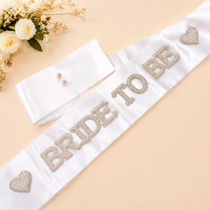May include: A white satin sash featuring the text BRIDE TO BE in silver-toned, rhinestone-encrusted letters. The sash is decorated with two heart-shaped rhinestone embellishments at the ends and includes a separate white fabric piece with two pins.