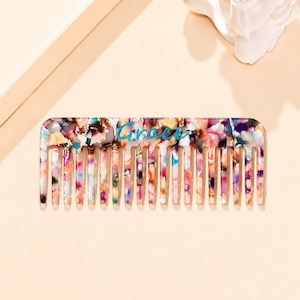 May include: A rectangular hair comb featuring a multi-colored, speckled pattern with shades of pink, blue, purple, and white. The word Grace is printed in a teal, cursive script across the center of the comb, which has numerous teeth.