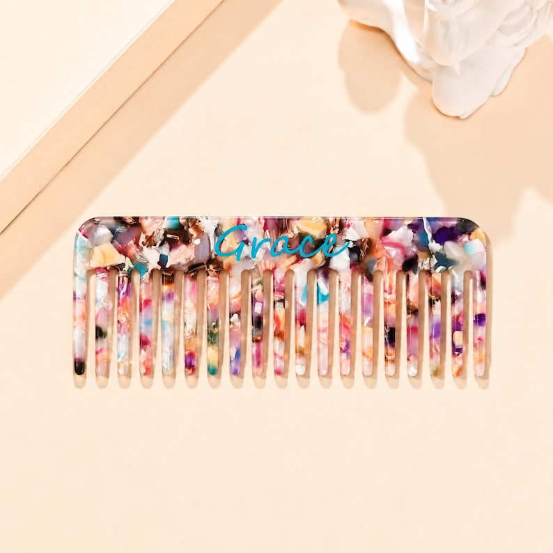 May include: A rectangular hair comb featuring a multi-colored, speckled pattern with shades of pink, blue, purple, and white. The word Grace is printed in a teal, cursive script across the center of the comb, which has numerous teeth.