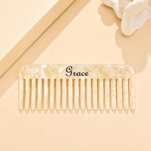 May include: A rectangular hair comb with a cream-colored pearlescent finish and a marbled texture. The word Grace is printed in a black script font across the center of the comb, which features a series of evenly spaced, vertical teeth.
