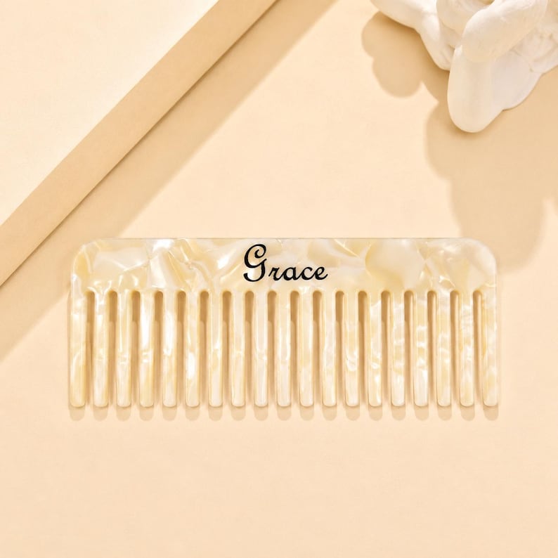 May include: A rectangular hair comb with a cream-colored pearlescent finish and a marbled texture. The word Grace is printed in a black script font across the center of the comb, which features a series of evenly spaced, vertical teeth.