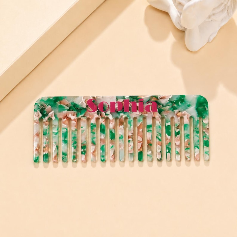 May include: A rectangular hair comb featuring a marbled pattern of green, white, and pink acrylic material. The center of the comb displays the name Sophia printed in a bold, dark pink font. The comb has evenly spaced teeth along the bottom edge.