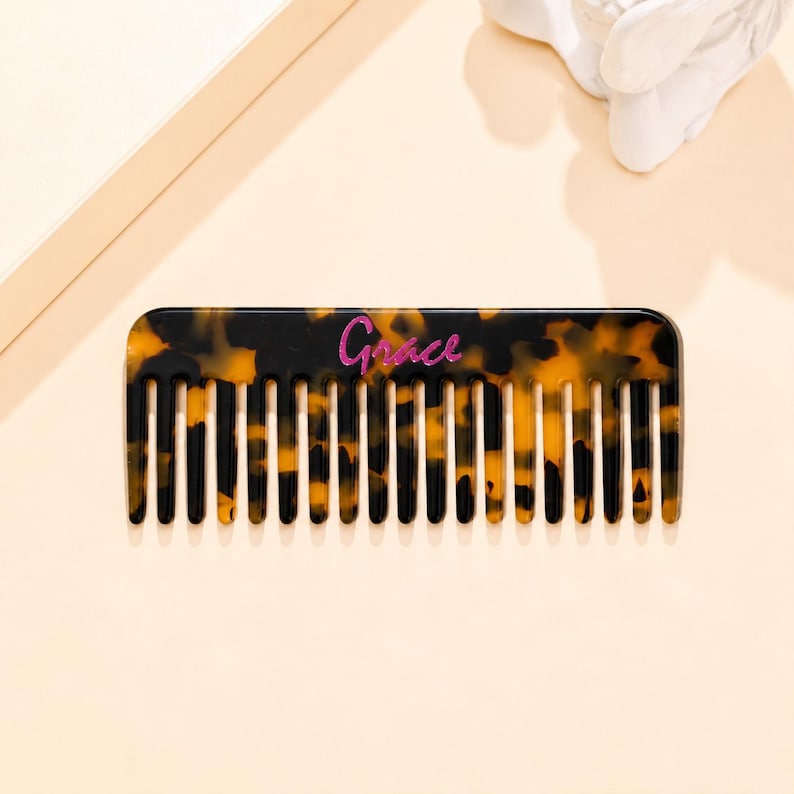 May include: A rectangular hair comb featuring a brown and black tortoiseshell pattern. The word Grace is printed in a cursive, glittery pink font across the center of the comb, which has wide, evenly spaced teeth along the bottom edge.
