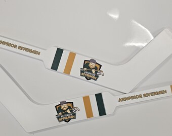 Puede incluir: Dos carteles blancos con forma de palo de hockey con las palabras "Arnprior Rivermen" y una imagen de dibujos animados de un jugador de hockey. Los carteles tienen rayas doradas, blancas y verdes.