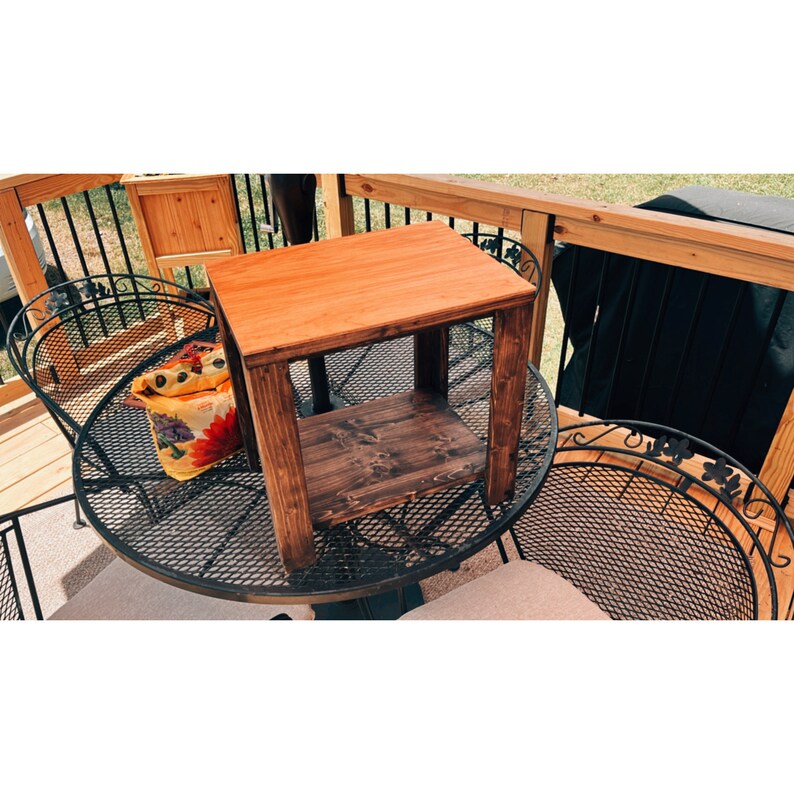 May include: A small wooden end table with a natural wood finish and a lower shelf. The table is sitting on a black metal patio table with a black metal chair next to it.