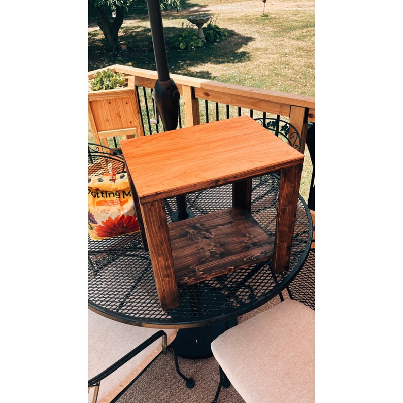 May include: A wooden end table with a light brown top and a dark brown bottom shelf. The table is sitting on a black metal patio table.