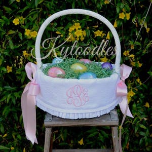 May include: White Easter basket with a handle, filled with green decorative grass and colorful Easter eggs. The basket has a pink monogram and is adorned with pink ribbons. The text "Kytoodles" is visible on the basket.