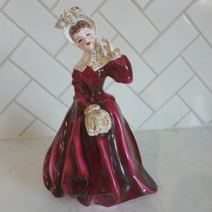 May include: A vintage porcelain figurine of a woman wearing a burgundy dress with a white lace collar and a white hat with gold accents. She is holding a bouquet of flowers in her hand.
