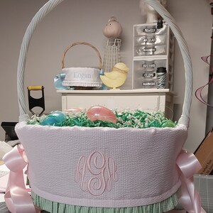 May include: A light pink Easter basket with a white handle and a monogram. The basket is filled with green paper grass and decorative eggs. Pink ribbons are tied on the sides. A smaller basket with the name "Logan" is in the background.