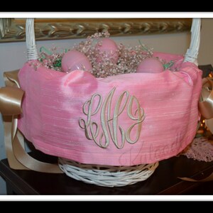 May include: A pink Easter basket with a white wicker base and a satin pink liner. The basket is decorated with a gold ribbon and a monogrammed "E.F.F." in gold thread. The basket is filled with pink and white Easter eggs and white baby's breath.